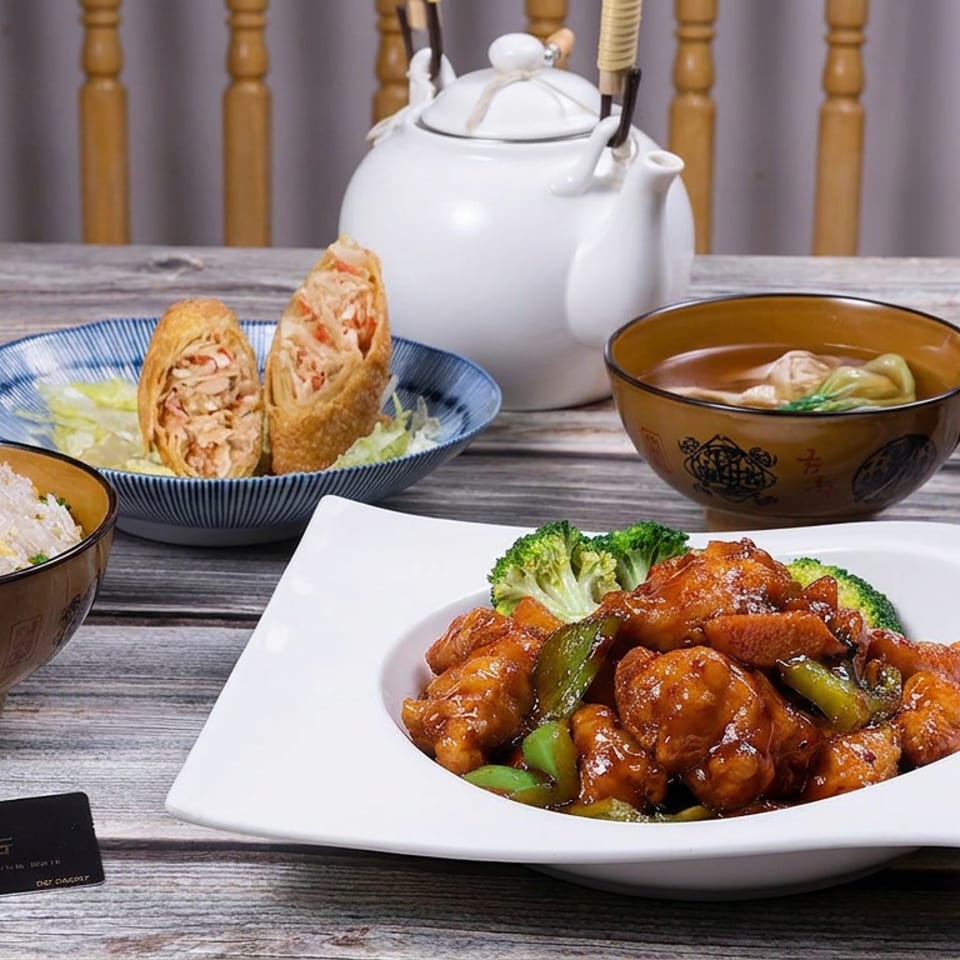 Chinese meal featuring glazed chicken with peppers and broccoli, fried rice, spring rolls, soup, teapot, and chopsticks on a rustic table.