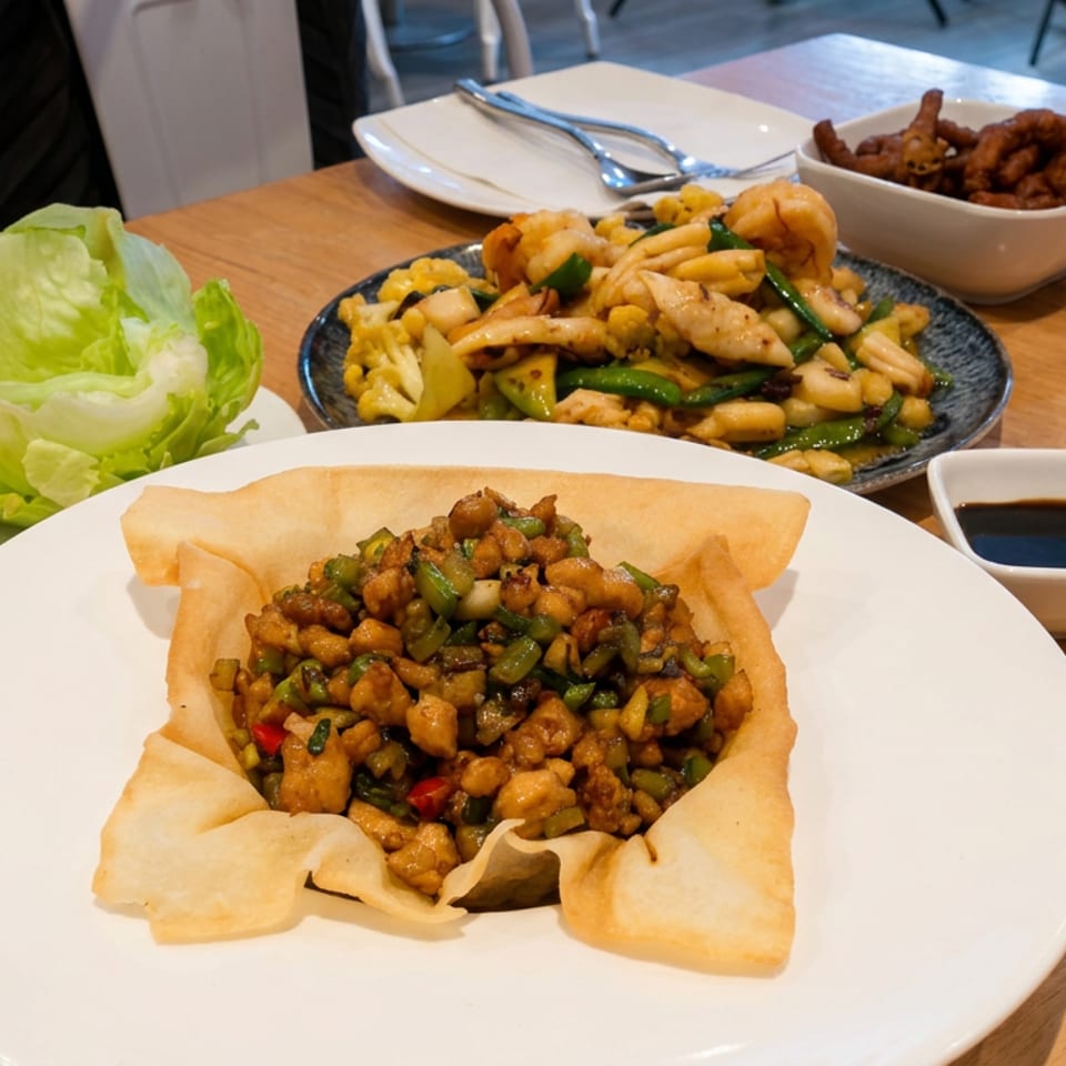 Chinese dishes fill a restaurant table, including crispy pastry with diced chicken, stir-fried seafood and vegetables, lettuce, and dipping sauce.