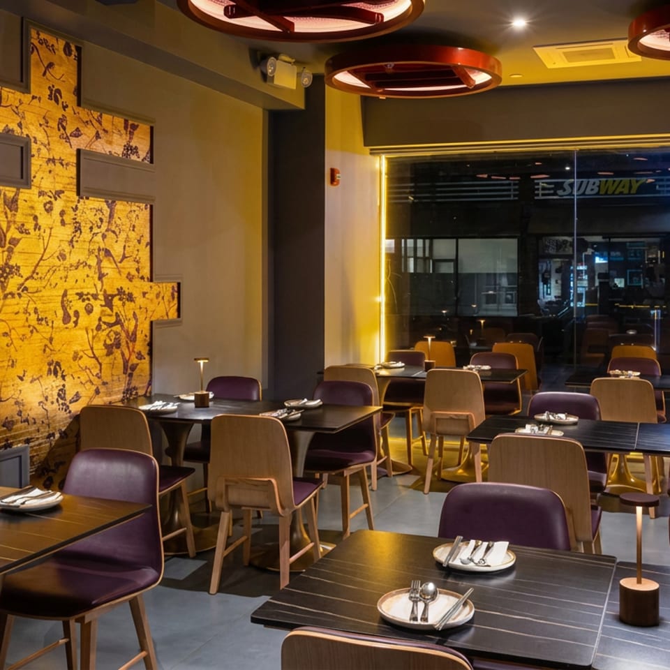 Warmly lit modern restaurant dining room with set tables, purple and wood chairs, patterned wall panel, and large windows.